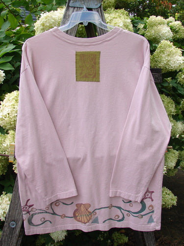 1993 Long Sleeved Tee Ocean Folly Ask Pink OSFA in a soft pink, long-sleeved shirt with a loose drape and a small square graphic at the upper back. A decorative sea-inspired border with shells and curling vine-like lines runs along the hem, adding muted teal and coral detail.