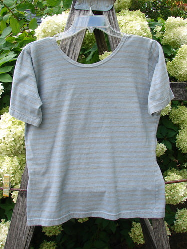 1997 Little Voyage Top in gray-blue stripes, a tiny size 2 T-shirt with a soft heathered knit texture, short sleeves, and a simple crew neckline. It hangs centered on a clear hanger against weathered wood and pale hydrangeas, with muted light softening the striped pattern.