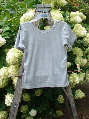 1997 Little Voyage Top Gray Blue Stripe Tiny Size 2, a small short-sleeve crewneck shirt in pale gray-blue with тонal horizontal stripes and a soft, lightweight knit finish. It hangs centered on a wooden ladder amid dense white hydrangea blooms and green leaves.
