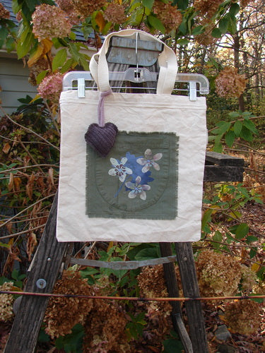 2000 PMU Patched Organic Cotton Canvas Pocket Tote Bag, featuring a purple heart and floral accents, displayed on a wooden ladder, showcasing its sustainable, vintage-inspired design from Bluefishfinder.com.