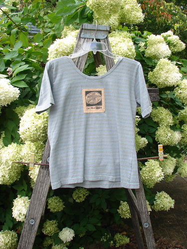 1997 Little Voyage Top Gray Blue Stripe Tiny Size 2, a short-sleeve top with fine gray-blue horizontal stripes and a centered square graphic patch. It hangs from a weathered wooden ladder amid dense white hydrangeas, with soft natural light and a muted, garden-toned palette.