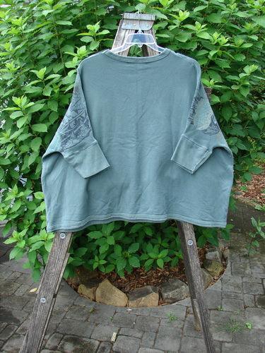 Barclay Fleece Banded Sleeve Colors Sweatshirt Moon Flower Olive Drab OSFA displayed on a wooden rack, showcasing its boxy shape, dolman banded sleeves, and moon flower theme against a rustic outdoor backdrop.