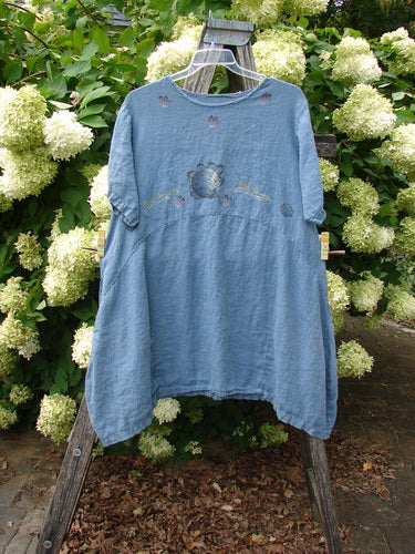 1997 Urchin Dress Floral Starfish Naya Size 1 in soft blue, a loose, short-sleeve dress with a gently gathered waist and a rounded neckline. The lightweight fabric shows a faded starfish motif, hanging from a clear hanger against white hydrangeas and greenery.