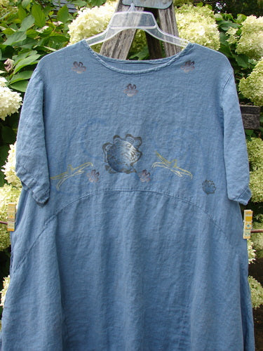 1997 Urchin Dress Floral Starfish Naya Size 1 in faded blue fabric, a loose short-sleeve dress with a rounded neckline, subtle floral and starfish motifs, and a softly gathered waist. It hangs on a clear hanger against leafy greenery and pale hydrangeas.