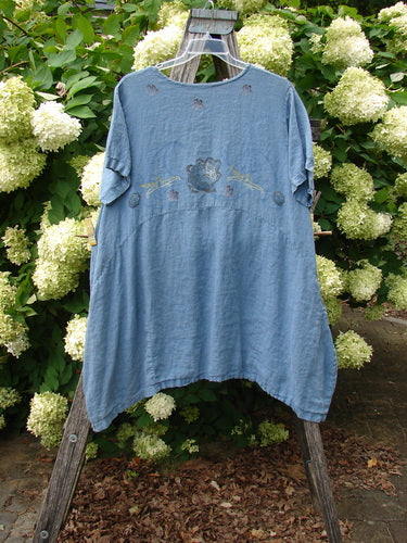 Naya 1997 Urchin Dress Floral Starfish Size 1 in faded blue, a loose short-sleeve dress with a soft, washed texture and subtle floral starfish motif across the chest. It hangs from a wooden ladder stand before dense white hydrangeas and green foliage, creating a rustic outdoor display.