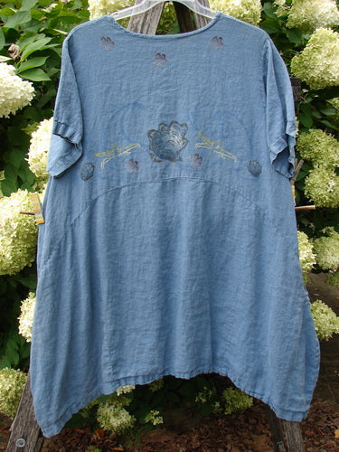 1997 Urchin Dress Floral Starfish Naya Size 1 in faded blue fabric, a loose short-sleeve dress with a round neckline, soft drape, and stitched floral starfish motifs across the chest. It hangs centered against leafy hydrangeas, with natural daylight emphasizing its worn texture.