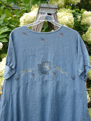 1997 Urchin Dress Floral Starfish Naya Size 1 in faded blue fabric, with a loose short-sleeve silhouette, rounded neckline, and embroidered floral and starfish motifs across the chest. It hangs on a clear hanger before leafy greenery and pale blossoms, with soft outdoor light deepening the washed, textured finish.