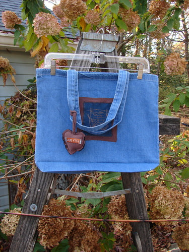 1998 PMU Patched Tote Bag Fall Quad Washed Denim One Size, featuring a colorful puffy heart accent, displayed on a ladder. The bag showcases vintage-inspired sustainable design with tote-length shoulder straps and gusset bottom.