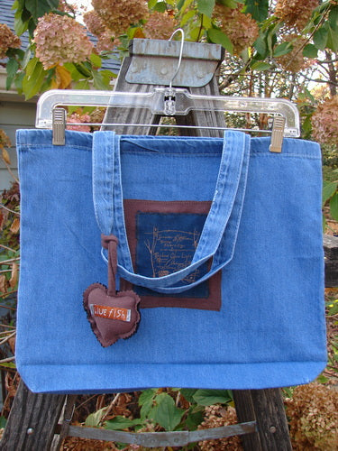 The 1998 PMU Patched Tote Bag Fall Quad Washed Denim One Size is displayed on a swing, featuring a colorful puffy heart accent, tote-length shoulder straps, and a gusseted bottom.