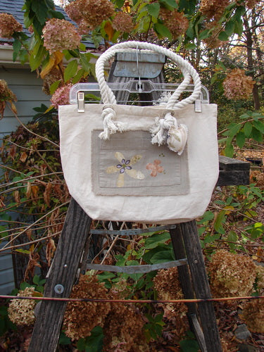 2000 PMU Patched Canvas Rope Pocket Tote Bag Daisy Natural One Size displayed on a ladder, featuring cotton rope shoulder straps, exterior pocket patches, and a signature Blue Fish patch, celebrating sustainable creativity.