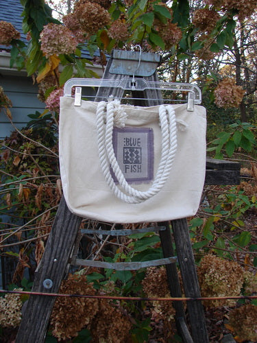 2000 PMU Patched Canvas Rope Pocket Tote Bag Daisy Natural One Size displayed on a wooden ladder, showcasing its cotton rope straps, front pocket patch, and signature Blue Fish label.