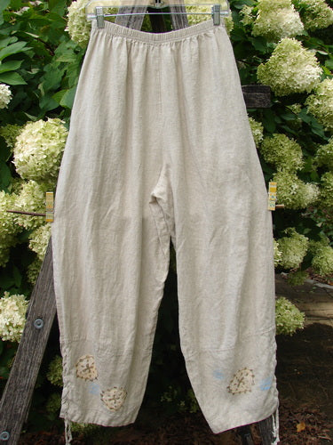 1997 Skipper Pant Rolling Sealife Natural Size 1 in soft natural linen, shown as loose, wide-leg pants with an elastic waistband and subtle rolled-sealife motifs near the hems. The pale beige fabric hangs in a relaxed drape against a leafy outdoor backdrop.