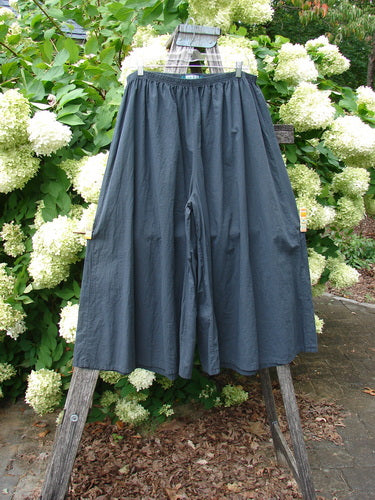 Barclay Batiste Crop Wide Leg Pant in unpainted black, size 2, with a softly crinkled matte finish and a loose cropped silhouette. The wide legs hang from a gathered waistband, photographed on a wooden ladder stand against dense white hydrangeas and green foliage.