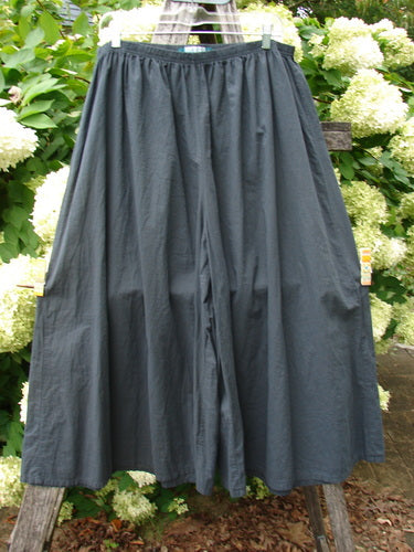 Barclay Batiste Crop Wide Leg Pant in unpainted black, size 2, with a gathered elastic waist and a flowing cropped wide-leg silhouette in matte, crinkled fabric. It hangs centered against leafy white hydrangeas and weathered wood, with soft natural light deepening the dark tone.