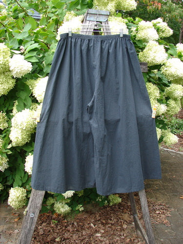 Barclay Batiste Crop Wide Leg Pant in unpainted black, size 2, with a voluminous cropped silhouette and wide straight legs. The matte woven fabric hangs from an elasticized waistband, shown clipped to a wooden rack against dense white blooms and green foliage.
