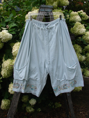 Barclay Bushel Short Planetarium Clear Blue Day Size 2 shorts in pale clear blue, with a relaxed wide-leg cut, elastic waistband, and rounded side pockets. Light fabric drapes softly, while a decorative planetary border traces the hem against a rustic wooden ladder and white hydrangeas.