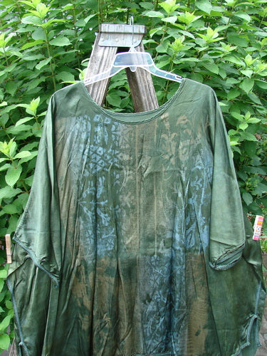 Barclay Viscose Vitreous High Low Tunic Botanicals Mottled Forest Sun OSFA displayed on a wooden pole, showcasing a rounded wider neckline, varying hem, and three-quarter dolman sleeves with exterior stitchery.