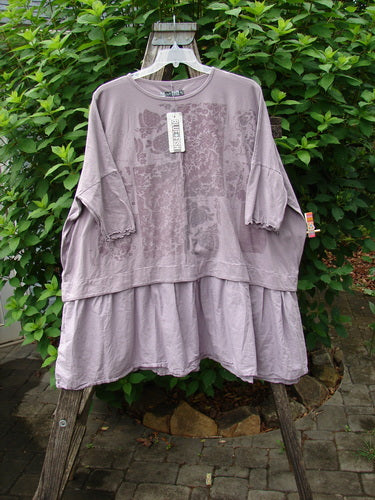 Barclay NWT Batiste Banded Flounce Esperanza Tunic Garden Sea Fog OSFA displayed outdoors on a hanger, showcasing its dolman sleeves, rounded neckline, and boxy shape with a floral theme.