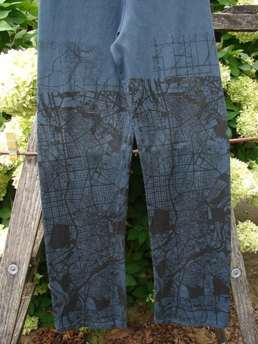 Barclay Cotton Lycra Straight Pant City Plan Lux Silhouette, size 0, in dark blue cotton-lycra with a black city-map print. The straight legs hang front and center, with a narrow slit at the hem, while the textured wood and greenery behind it frame the pants in soft daylight.