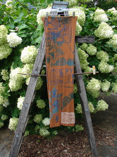 Barclay Curly Edge Center Seam Scarf Wrap in Fall Forest Friend rust, a long rectangular wrap with a curly edge and center seam. The sheer, lightweight fabric shows layered blue forest motifs and a small pale patch near the hem, hung vertically against greenery.