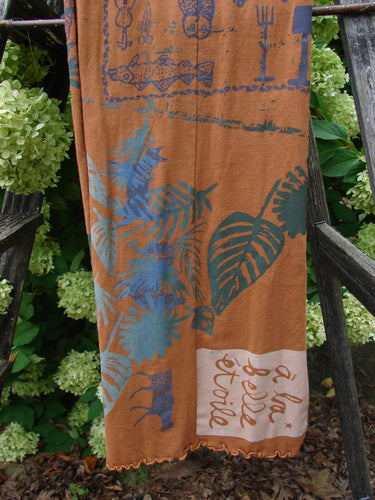 Barclay Curly Edge Center Seam Scarf Wrap in rust, a long draped scarf with a curly scalloped edge and center seam. Its soft fabric shows layered teal and blue botanical and fish motifs, plus a pale corner panel with script, hanging against leafy greenery and dark wood.