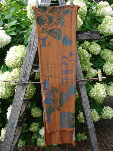 Barclay Curly Edge Center Seam Scarf Wrap in fall forest friend rust, a long rust-brown wrap with blue leaf and bird motifs, a center seam, and a softly curled edge. It hangs vertically against a weathered wooden ladder, with pale hydrangea blooms behind it.
