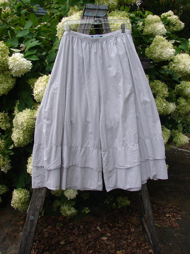 Barclay Batiste Cotton Wide Leg Carassai Pant in unpainted stone, size 2, shown as a light gray cotton skirt-like pant with a gathered elastic waist and wide, flowing legs. The soft matte fabric hangs from a wooden ladder amid white hydrangeas and dark greenery.