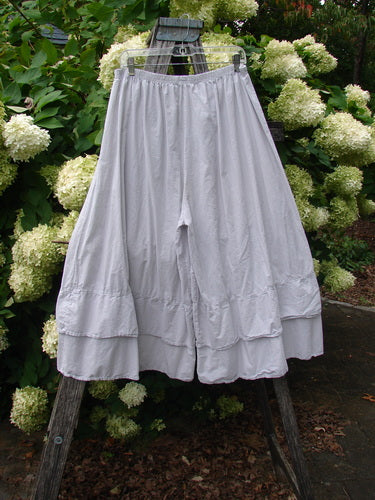 Barclay Batiste Cotton Wide Leg Carassai Pant in unpainted stone, size 2, shown as a full-length wide-leg skirted pant with a soft gathered waistband and layered hem. The lightweight cotton fabric hangs in a pale matte drape against green hydrangea blooms.
