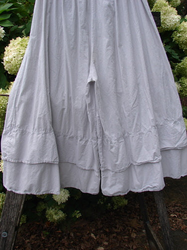 Barclay Batiste Cotton Wide Leg Carassai Pant in unpainted stone, size 2, with a soft cotton texture and sweeping wide legs finished by a layered hem. The light gray fabric hangs in deep vertical folds, framed by outdoor greenery.