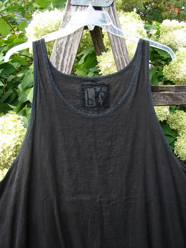 Barclay Linen Pinafore Curve Drop Side Rose Border Black Size 2, a black linen pinafore with a soft A-line drape and curved drop sides, hanging on a clear hanger against weathered wood and pale hydrangea blooms.