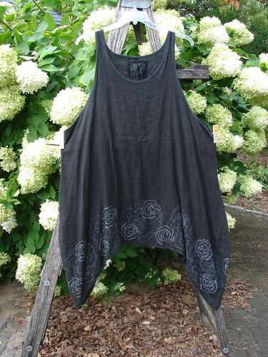 Barclay Linen Pinafore Curve Drop Side Rose Border Black Size 2, a sleeveless black linen pinafore with a loose, curved silhouette and deep drop sides. Pale rose border motifs trace the lower hem, while the garment hangs from a wooden ladder against dense white blossoms and greenery.