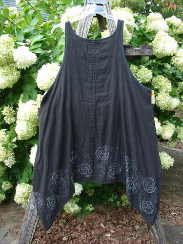 Barclay Linen Pinafore Curve Drop Side Rose Border Black Size 2, a sleeveless black linen pinafore with a loose A-line drape and curved dropped sides. Pale rose-border spiral motifs trace the hem, while the garment hangs from a wooden ladder among white hydrangeas and green foliage.