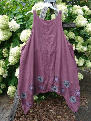 Barclay Linen Flared Pinafore Shift Dress in berry size 2, with a sleeveless A-line silhouette and uneven pointed hem. The linen fabric has a soft, textured drape, and circular pinwheel turn leaf motifs cluster along the lower edge against a backdrop of white hydrangeas and greenery.