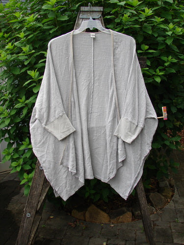 Barclay Gauze Open Front Cardigan Unpainted Natural Size 2 displayed on a wooden stand, showcasing its generous dolman sleeves, fully open front, and contrasting lower cuffs with a varying hemline.