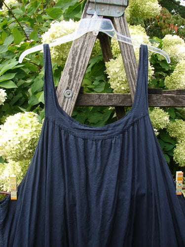 Barclay Batiste Banded Collar Slip Dress Birdy Border in black, size 2, hangs from a clear hanger with slim straps and a softly gathered neckline. The dark fabric falls in a smooth, loose drape against leafy hydrangeas and weathered wood.