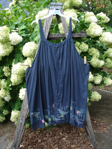 Barclay Batiste Banded Collar Slip Dress Birdy Border in black, size 2, hangs from a weathered wooden stand with thin shoulder straps and a loose, gathered silhouette. The lightweight fabric falls in a soft drape, finished with a subtle bird-and-border pattern near the hem, against lush green hydrangeas.