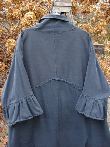 A woman wearing a Barclay Decora Brushed Twill Flutter Coat in Navy, featuring a deep V-shaped neckline, dark matching buttons, and flutter ruffle 3/4 length sleeves. The coat has a wrap-around rear with an upward pointed waistline, giant drop round bottom pockets, and a paneled and widening swingy hemline. Made from a heavy brushed cotton twill, this coat is a perfect addition to your wardrobe.