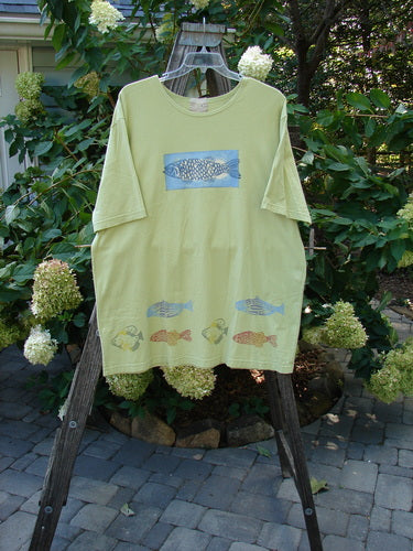 2000 Short Sleeved Tee Fish School Key Lime Pie Size 2 displayed on a swinger, showcasing its medium weight organic cotton fabric and fish school design with drop shoulders and wider cut sleeves.