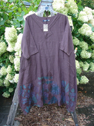 Barclay NWT Linen Cross Over Incense Dress in fig, size 0, with a loose V-neck crossover bodice, elbow-length sleeves, and a soft linen texture. Blue floral and diamond motifs cluster along the hem against a garden backdrop.