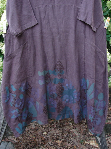 Barclay NWT Linen Cross Over Incense Dress Structural Diamond Floral Fig, size 0, in muted plum linen with a draped crossover bodice and loose short sleeves. The hem falls in soft folds over a darker floral-and-diamond print, creating a layered, airy silhouette.