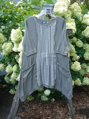Barclay Raw Edge Flutter Drop Pocket Tunic Woods in darkest sand, size 0, with a soft draped silhouette, raw-edge seams, flutter sleeves, and drop pockets. The tunic hangs from a wooden display amid white hydrangeas, its muted gray-green fabric and textured front panel catching soft daylight.