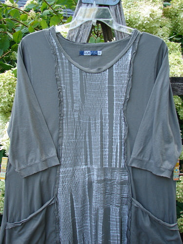 Barclay Raw Edge Flutter Drop Pocket Tunic in Woods Darkest Sand, size 0, with a soft dark gray body and a patterned front panel. The tunic shows raw-edged flutter trim, three-quarter sleeves, and curved drop pockets, hanging on a hanger outdoors.