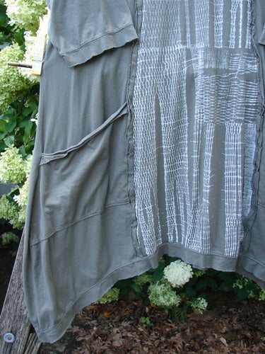 Barclay Raw Edge Flutter Drop Pocket Tunic in darkest sand, size 0, with a soft draped silhouette, raw-edged flutter layers, and a loose side pocket. The muted fabric shows a textured panel with pale woven lines, hanging in layered folds against greenery.