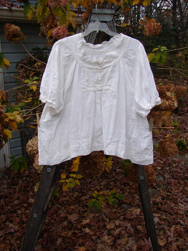 Magnolia Pearl Homespun Linen Lace Button Bib Top, displayed on a clothes rack, showcasing its three-quarter sleeves, lace accents, and unique hemline.