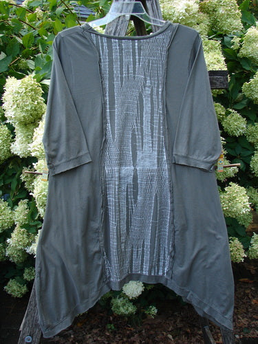 Barclay Raw Edge Flutter Drop Pocket Tunic in Woods Darkest Sand, size 0, with a dark sand body, raw edges, flutter sleeves, and a long front panel in a pale textured grid pattern. The tunic hangs on a clear hanger, its soft drape and uneven hem set against dense green foliage and white blossoms.