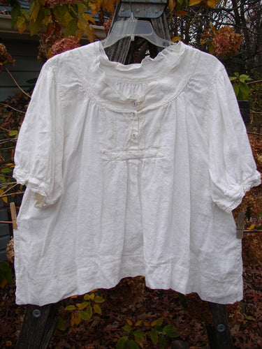 Magnolia Pearl Homespun Linen Lace Button Bib Top Farm Love Off White OSFA hangs on a clothesline, showcasing its three-quarter sleeves, lace accents, and distinctive Magnolia Pearl rear patch.