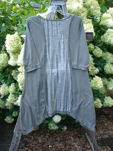 Barclay Raw Edge Flutter Drop Pocket Tunic in Woods Darkest Sand, size 0, shown as a loose gray-green tunic with raw edges, elbow-length flutter sleeves, and a textured center panel. The soft drape and uneven hem create a layered silhouette against dense green hydrangeas.