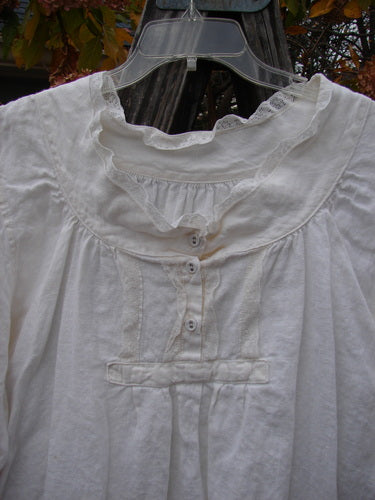 Magnolia Pearl Homespun Linen Lace Button Bib Top, displayed on a hanger, showcases lace accents on the bib, cuffs, and neckline, with three-quarter sleeves and a varying hemline.