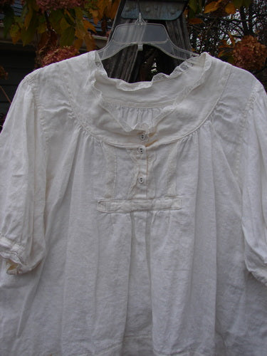 Magnolia Pearl Homespun Linen Lace Button Bib Top Farm Love Off White OSFA displayed on a hanger, showcasing its three-quarter sleeves, lace accents, and unique hemline.