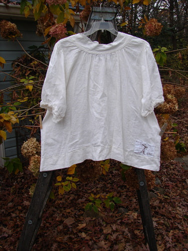 Magnolia Pearl Homespun Linen Lace Button Bib Top Farm Love Off White OSFA displayed on a swing, showcasing its three-quarter sleeves, lace bib accents, and varying hemline in an outdoor setting.